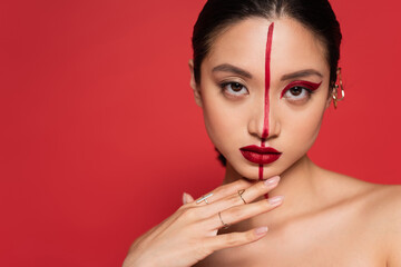 portrait of asian woman with creative visage touching chin and looking at camera isolated on red.