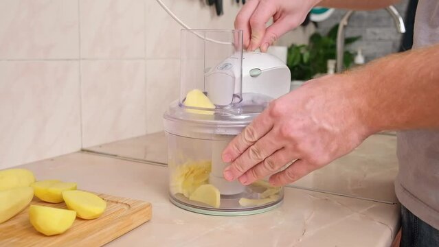 A man cuts potatoes into thin slices in a food processor. Preparation of potatoes for cooking. A man cooks dinner or lunch at home for the family. Kitchen appliances.