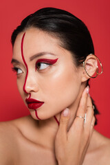 portrait of asian woman with creative visage and ear cuff touching neck and looking away isolated on red.
