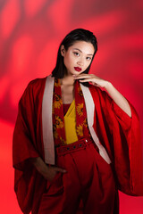 Fototapeta na wymiar elegant asian woman in kimono cape and scarf with floral print standing with hand in pocket on red shaded background.