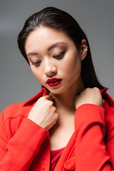 portrait of brunette asian woman with makeup touching collar of red jacket isolated on grey.