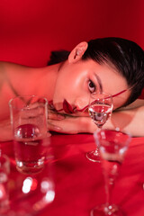 brunette asian woman with artistic makeup lying and looking at camera near blurred glasses on red background.