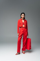 full length of asian model in red jacket and trousers standing with shopping bag on grey background.