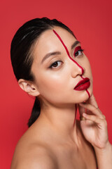 Fototapeta premium portrait of asian model with creative visage and perfect skin holding hand near face and looking at camera isolated on red.