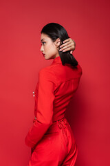 side view of brunette asian woman in stylish suit fixing hair while looking away on red background.