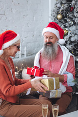 Fototapeta premium Happy loving senior couple having fun on New Year party near Christmas tree. Mature man and woman smiling, drinking champaign, exchanging gifts. Concept of active life and leisure of pensioner