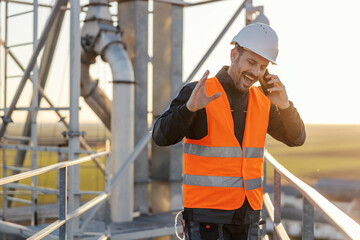 An industry worker is standing on the high metal construction and talking on the phone with manager.