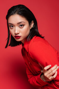 portrait of brunette asian woman with makeup and ear cuff looking at camera on red background.