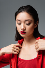 young asian woman with red lips touching stylish jacket isolated on grey.