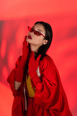 sensual asian woman in gloves and sunglasses looking at camera on coral red background with shadow.