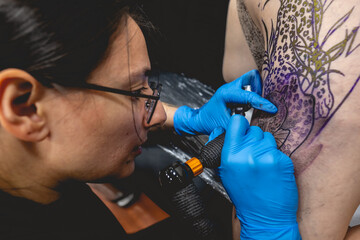 Young tattoo artist girl with glasses making a tattoo, with tattoo machine, of a big tiger, in the...