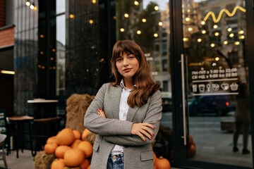 Pretty adorable dark-haired woman wearing gray jacket and white blouse posing at camera over city holiday background