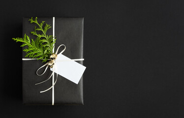 Black gift box with sprig of thuja, bells and tag on black background. Space for your text.