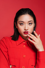 portrait of pretty asian woman in stylish jacket looking at camera and touching lips isolated on red.