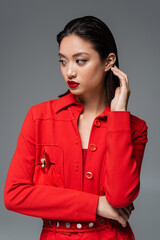 fashionable asian woman in red blazer touching brunette hair and looking away isolated on grey.