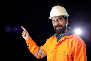 Man with beard wearing hardhat and reflecting jacket smiling, looking at the camera pointing finger to the side.