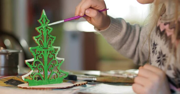 The holiday season. The hands of a small child coloring a toy wooden Christmas tree with a brush. Hand held. Close-up.