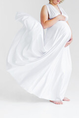 A pregnant woman in a long white flying silk dress hugs her belly and poses on a white background