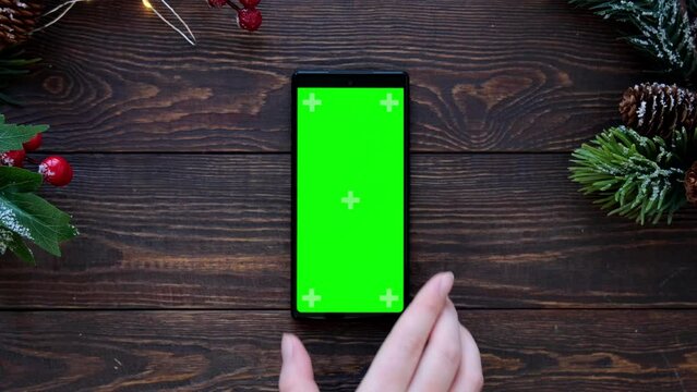 Women's hands puts the phone with a Green screen on a wooden table with christmas decor. Top view of smartphone 