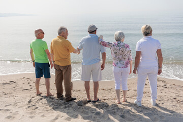 gruppo di 5 anziani in spiaggia in piedi  guardano abbracciati  l'orizzonte in lontananza 