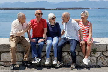 gruppo di anziani amici seduti nel muretto di un porto di mare, si rilassano chiacchierando felici.