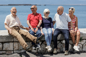 gruppo di anziani amici seduti nel muretto di un porto di mare, si rilassano chiacchierando felici. - obrazy, fototapety, plakaty