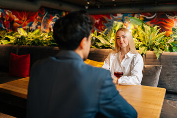 View from back of unrecognizable young man to beautiful blonde woman sitting at table with glass of red wine having festive dinner at restaurant. Happy loving couple enjoying talking together.