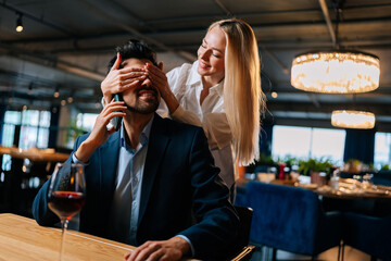 Joyful blonde young woman closed eyes of surprised elegant man in suit sitting at table with glass red wine talking on mobile phone. Happy beautiful female came on date with smiling male.