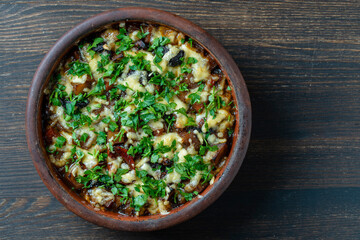 Stewed potato, carrot, onion, tomato, prunes and cheese in a clay bowl on wooden background, closeup, top view