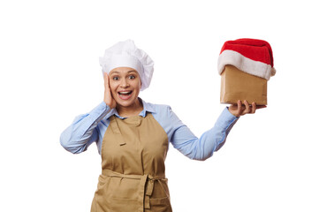 Delightful woman, chef confectioner in apron, expressing astonishment looking at camera, holding a paper bag with Santa hat, isolated over white background with copy space. Food delivery at Christmas