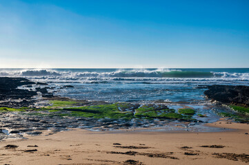 A powerful ocean wave crashes and breaks, creating foamy white spray as it approaches a rocky shoreline.