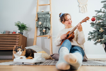 Young woman playing ukulele guitar while relaxing with cat pet on floor cushions near potted...