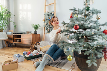 Young woman in cozy sweater decorating potted Christmas tree while her cat is watching her in light modern Scandinavian interior. Eco-friendly winter holidays. Domestic pet at home. Selective focus.
