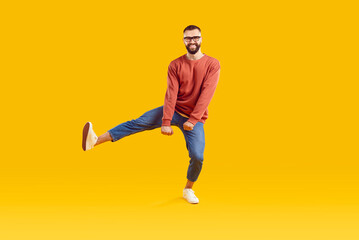Fototapeta na wymiar Smiling funky cheerful young bearded man with short hair in glasses wearing red sweatshirt and denim pants dancing on one leg isolated on yellow background. Good mood happy lifestyle nice day concept.