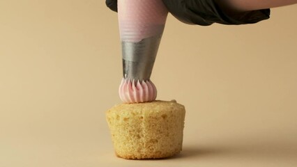 Close-up of confectioner squeezing whipped pink buttercream out of the pastry bag on top of the muffin. Pastry chef making cupcake in a slow motion on the warm beige background. Happy Valentine's day