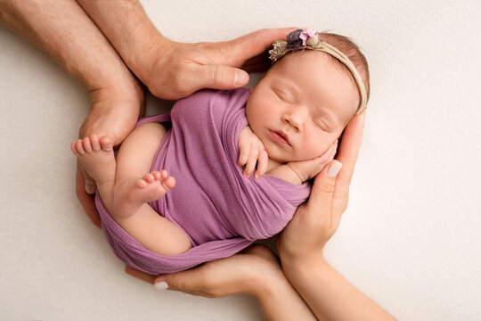 Sleeping newborn girl in the first days of life on a white background. A newborn baby in a purple lilac winding and a headband. Hand, palms of father and mother, parents hold the child.