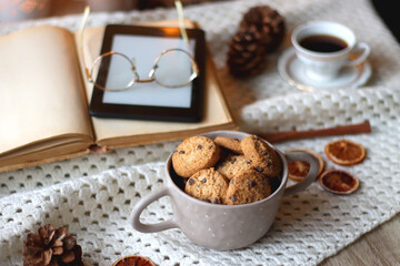 Cup of tea, books, tablet, bowl of cookies, various spices, pine cones and lit candles. Hygge at home. Selective focus.