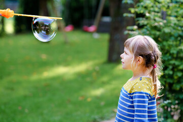 Happy little blonde preschool girl having fun with blowing soap bubble blower. Cute child playing...