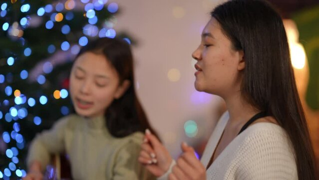Side view joyful Asian woman singing Christmas carol with blurred friend playing guitar at background at New Year tree. Happy confident positive ladies enjoying holiday celebration indoors
