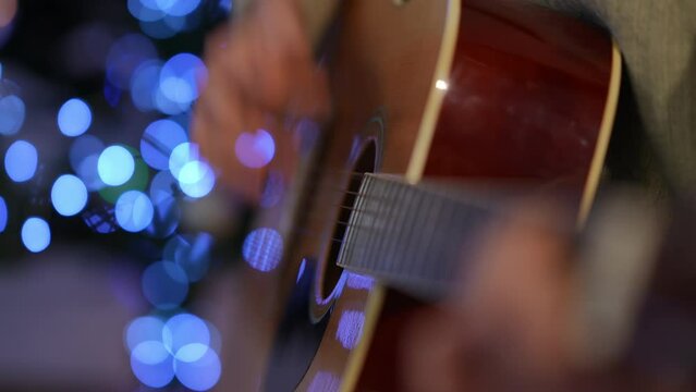 Close-up guitar fretboard with slim female hand playing strings at background of Christmas light. Unrecognizable talented woman enjoying hobby singing New Year carol sitting indoors
