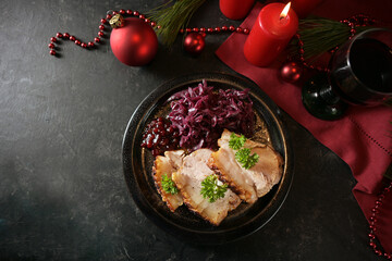 Festive dinner, roast pork slices with red cabbage on a dark plate, red candles, wine and Christmas decoration on a dark background, high angle view from above, copy space