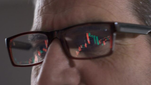 Trader analyzing charts, trading cryptocurrencies on online stock market.
Graphics reflected in the glasses of the man watching the stock market on the computer. Careful tracking and green, red graphi