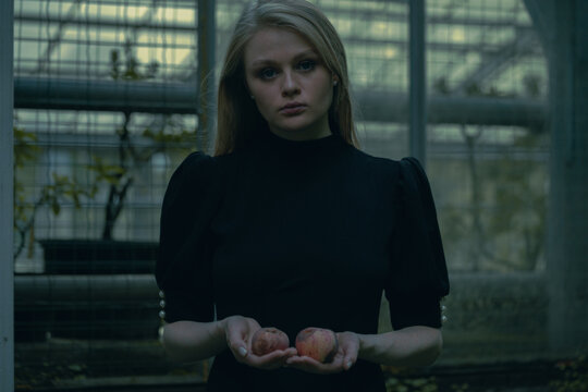 Beautiful Blond Girl Holding Rotted Wormy Apples Near Abandoned Houseplant At Gothic Park. Concept Of Doubts And Choice