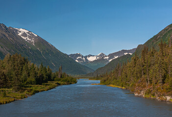 Fototapeta na wymiar Moose Pass, Alaska, USA - July 22, 2011: Blue water Clear Creek meandering between green forests and dark mountains with snow patches under blue sky