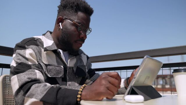 black man with glasses is surfing internet by tablet with free wifi in city cafe, working online