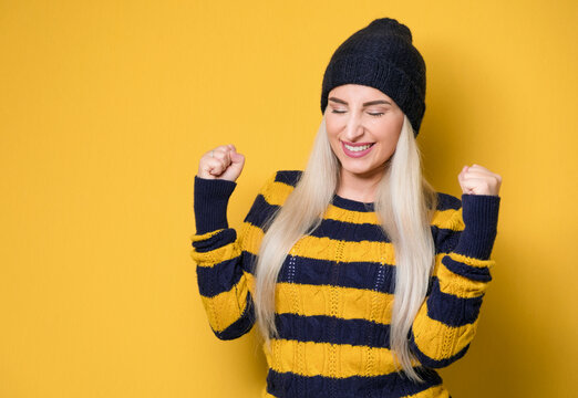 Excited woman with clenched fists and teeth, model wearing woolen cap and sweater, isolated on yellow background. Yes concept. Good news. Rejoicing female celebrates