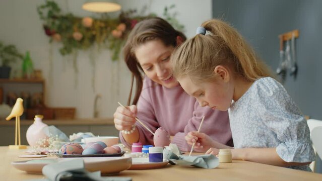 Little girl painting with cheerful mother while making Easter decorations at home