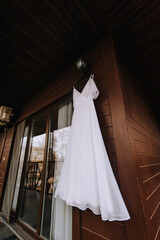 A beautiful long white lace bride's dress hangs on a hanger in the morning against the background of a wooden wall in the house. Wedding photography.
