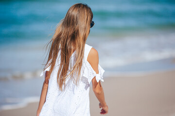 Fototapeta na wymiar Back view of adorable little girl with long hair in white dress walking on tropical beach vacation