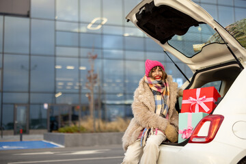 Young cheerful woman sits at car trunk full of presents near mall outdoors. Concept of winter...